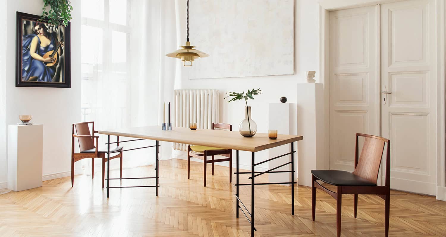 Sunny and elegant dining room interior with design sharing table, chairs, gold pedant lamp, abstract paintings and stylish accessories. Tropical leafs in vase. Eclectic decor. Brown wooden parquet.